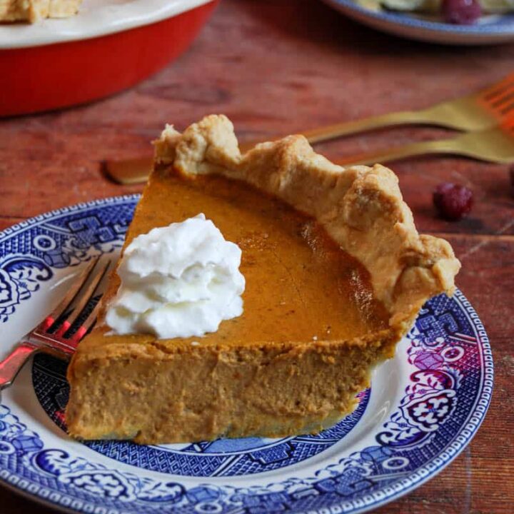 A slice of pumpkin pie topped with whipped cream on a blue willow pattern plate with a fork. A large pumpkin pie and another slice of pie on a plate can be partially seen in the background.