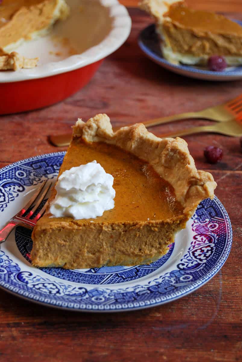 A slice of pumpkin pie topped with whipped cream on a blue willow pattern plate with a fork. A large pumpkin pie and another slice of pie on a plate can be partially seen in the background.