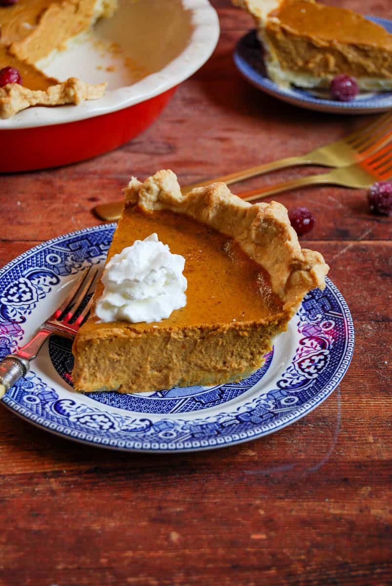 A slice of pumpkin pie topped with whipped cream on a blue willow pattern plate with a silver fork.
