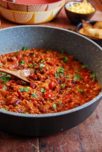 A grey pan of turkey chilli sprinkled with fresh coriander. Yellow and orange bowls, a pot of grated cheese and a bowl of fried tortilla chips are in the background.
