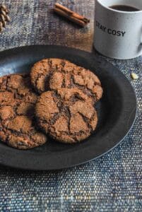 Five gingerbread cookies on a black plate, a white mug of coffee with the words "stay cosy" printed on the front and a cinnamon stick.