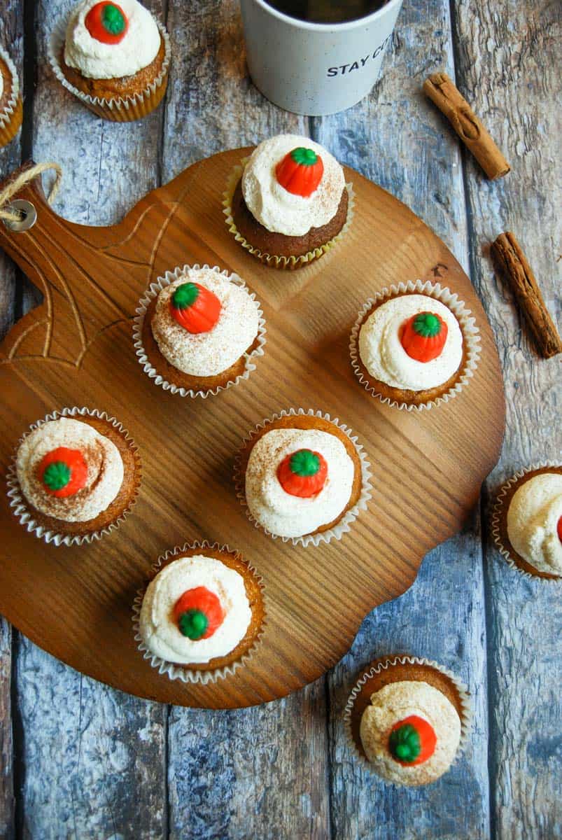 Six pumpkin spice latte cupcakes topped with whipped cream and a fondant pumpkin on a wooden pumpkin serving board. A mug of coffee, cinnamon sticks and more cupcakes surround the board.