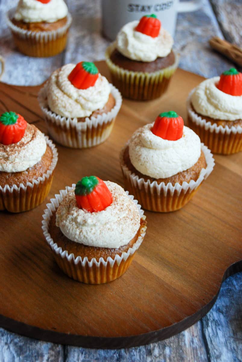 Six pumpkin spice cupcakes topped with whipped cream and a fondant pumpkin on a wooden board.