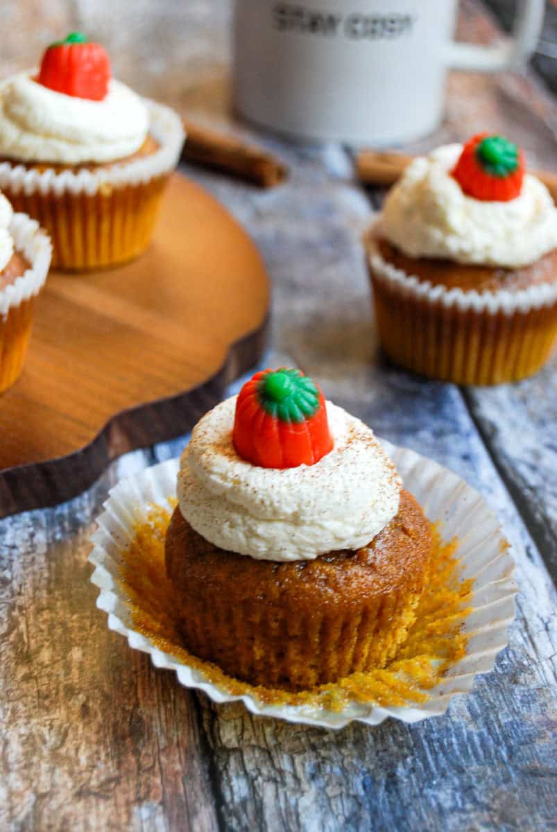 A pumpkin spice latte cupcake topped with whipped cream and a fondant pumpkin. The cupcake wrapper has been peeled back to reveal the orange colour of the sponge. More cupcakes, a white mug and cinnamon sticks are in the background.