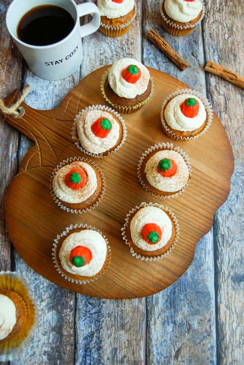 Seven Pumpkin spice latte cupcakes topped with whipped cream, dusted with ground cinnamon and topped with a fondant pumpkin on a wooden pumpkin serving board. A cup of coffee, more cupcakes and cinnamon sticks surround the board.