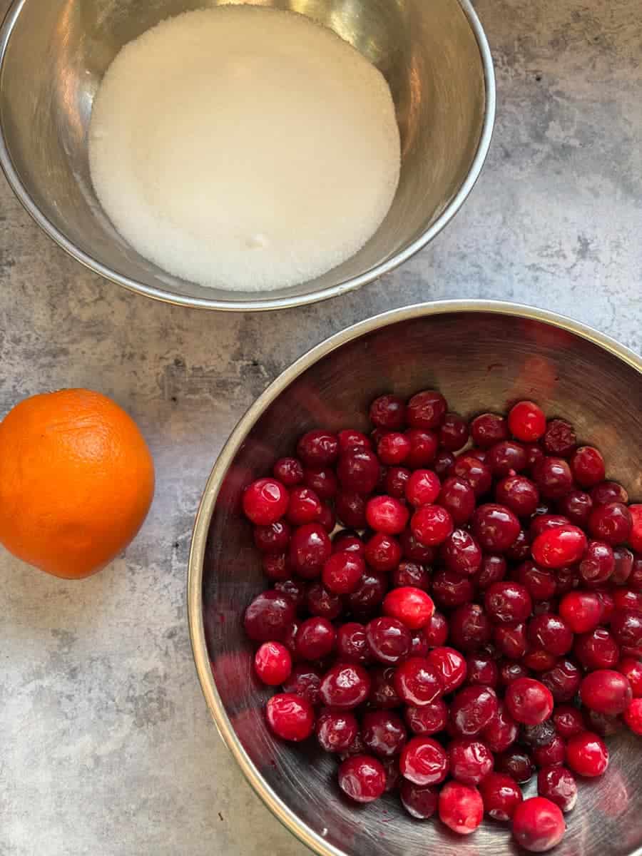 A silver bowl of fresh cranberries, a silver bowl of white sugar and an orange on a blue-grey surface.