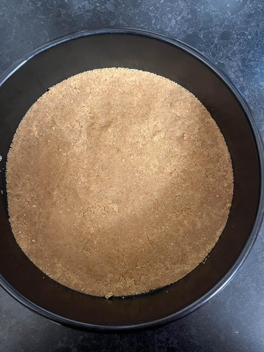 A round black cake tin filled with a digestive biscuit crust for a cheesecake.