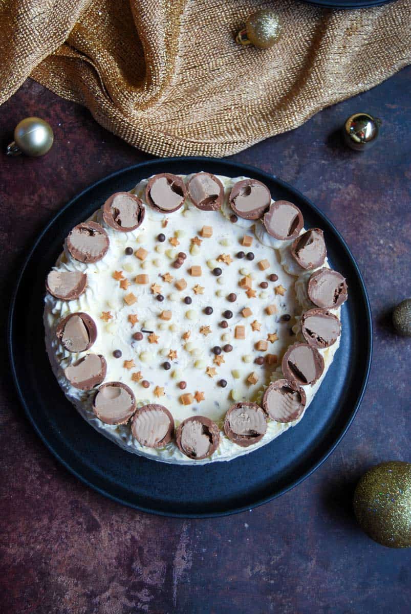 A Baileys cheesecake decorated with Irish cream filled chocolate truffles and gold star decorations on a black plate. A gold tablecloth and gold baubles are placed around the cheesecake.