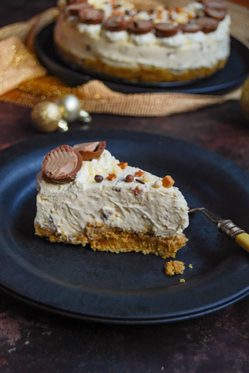 A slice of Baileys cheesecake decorated with Irish cream filled chocolate truffles and small gold stars on a black plate with a yellow fork. A large cheesecake sits on a black plate in the background with a gold scarf and gold Christmas baubles.