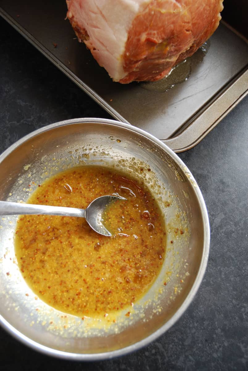 A silver bowl of orange marmalade glaze and a large cooked ham on a silver tray.