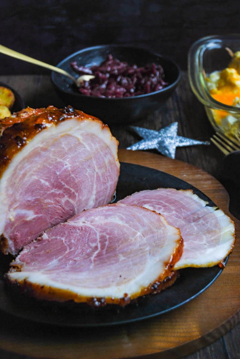 A partially sliced cooked ham on a black plate, a black bowl of red cabbage with a gold spoon, a dish of cauliflower cheese and a silver star decoration.