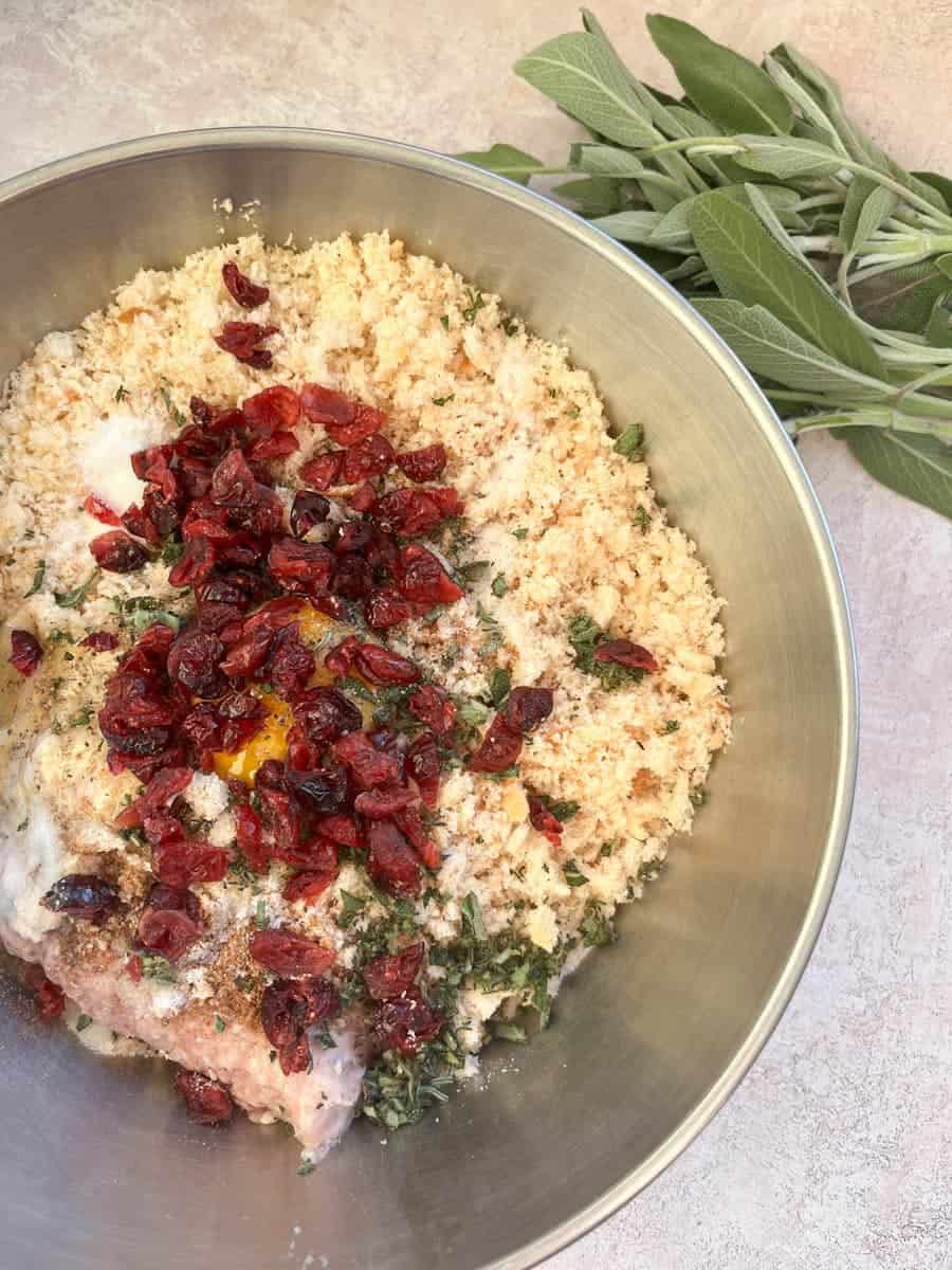 A silver bowl of sausage meat, breadcrumbs, fresh chopped sage, dried cranberries and an egg to make stuffing. Fresh sage leaves have been placed beside the bowl.