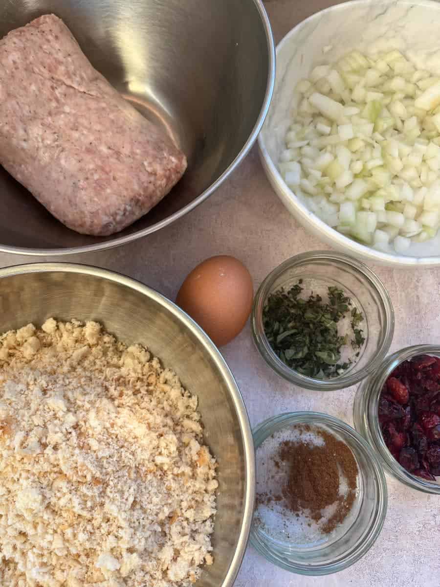 Silver bowls of sausage meat, breadcrumbs, a white bowl of chopped onion, an egg and glass jars of dried cranberries, mixed spice and chopped fresh sage.