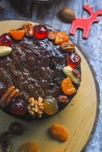 A small round Christmas fruit cake glazed with apricot jam and decorated with glazed pecans, almonds, walnuts, brazils, glace cherries, apricots and stem ginger on a wooden serving board. A wooden red Reindeer Christmas decoration and mixed nuts and fruit are placed beside the cake for a festive theme.