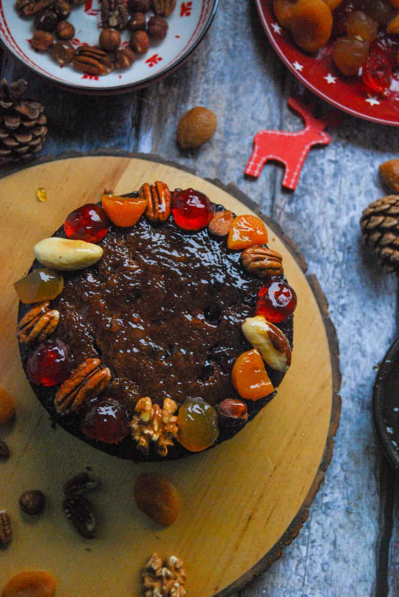 A small round Christmas cake glazed with dried fruits and nuts - glace cherries, stem ginger, dried apricots, pecans, walnuts and almonds on a wooden serving board. A small red plate of dried fruits and a red and white bowl of mixed nuts can be partially seen. A red wooden reindeer decoration, nuts and pine cones have been placed beside the board.