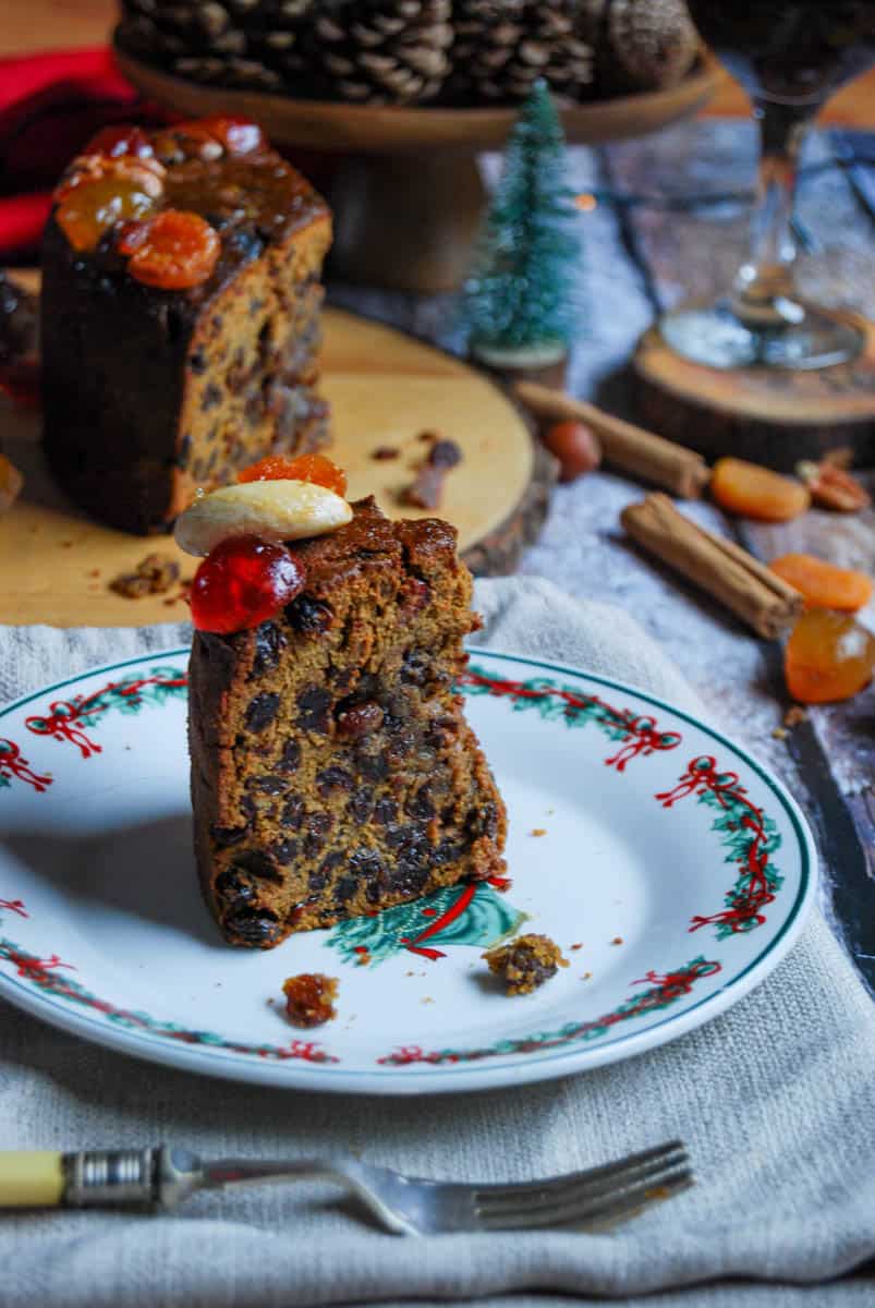 A slice of Christmas cake topped with a glace cherry and a brazil nut on a Christmas themed plate with a yellow handled fork. A Christmas cake that has been sliced sits in the background on a wooden board with a glass of mulled wine, cinnamon sticks, dried apricots and nuts, a tiny fir tree and a wooden cake stand with pine cones.