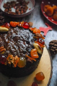A round fruit cake glazed with apricot jam and decorated with whole nuts and dried fruits on a wooden board.