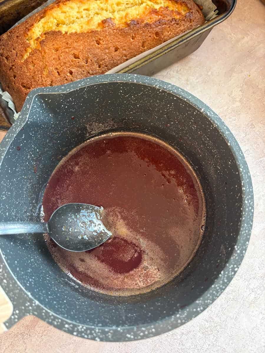 A grey pan of blood orange syrup with a silver spoon and an orange loaf cake.