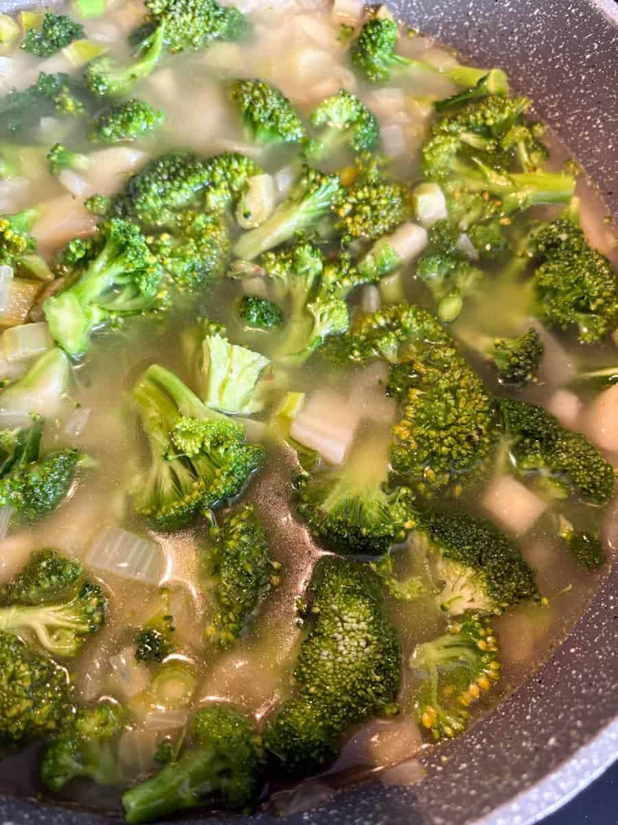 Chopped broccoli in a grey pan with stock, diced onion, potatoes and celery to make Broccoli soup.