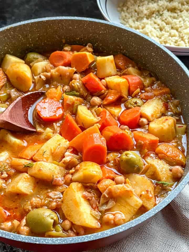A grey pan of vegetable stew with diced carrots, parsnips, sweet potatoes, leeks, chickpeas and green olives and a wooden spoon. A bowl of couscous can be partially seen above the pan.