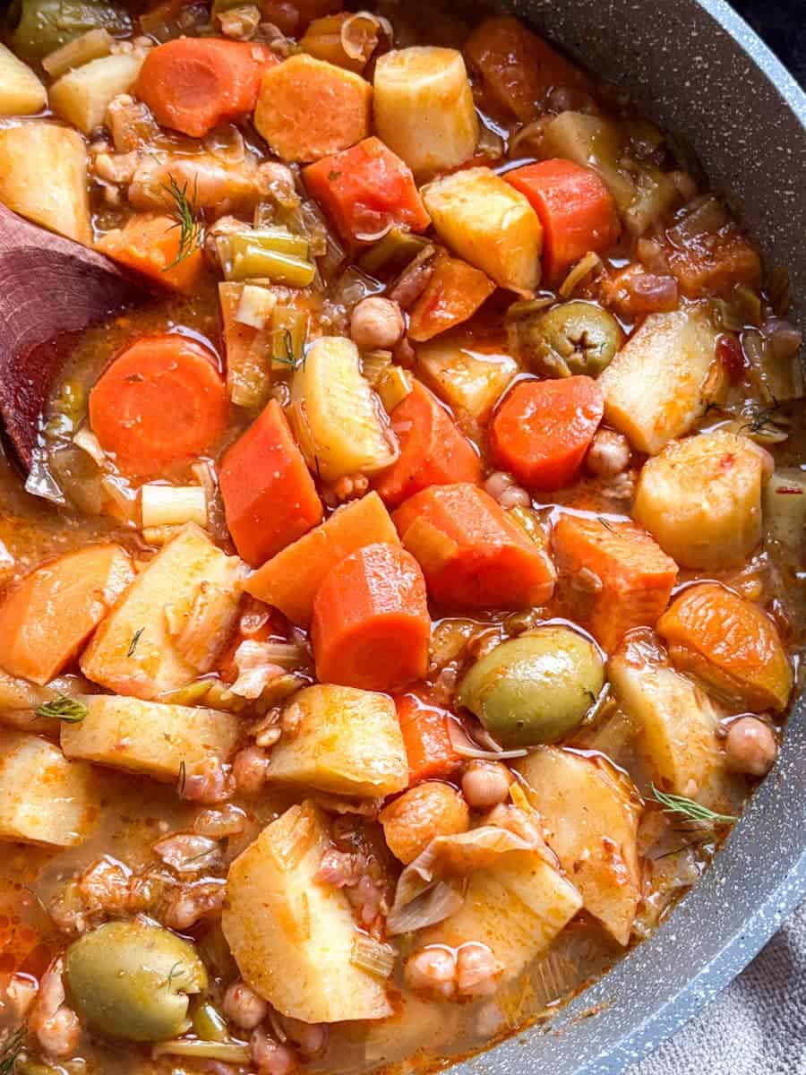 A close up image of a pan of vegetable stew with diced carrots, parsnips, sweet potatoes, leeks, chickepeas and green olives.