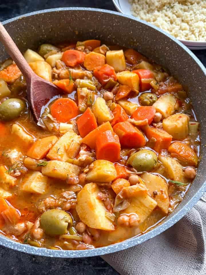 A pan of vegetable stew with diced carrots, parsnips, leeks, sweet potatoes, green olives and chickpeas with a wooden spoon. A white bowl of couscous can be partially seen.