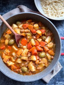 A grey and red pan of vegetable stew with sliced carrots, parsnips, sweet potatoes, sliced leeks, chickpeas and green olives with a wooden spoon and a white bowl of couscous.