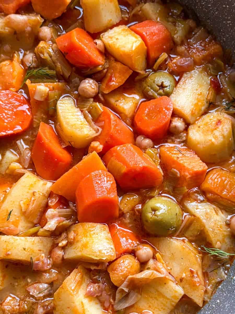 A close up image of a pan of vegetable stew with chopped carrots, parsnips, sweet potatoes, chickpeas and green olives in vegetable broth.