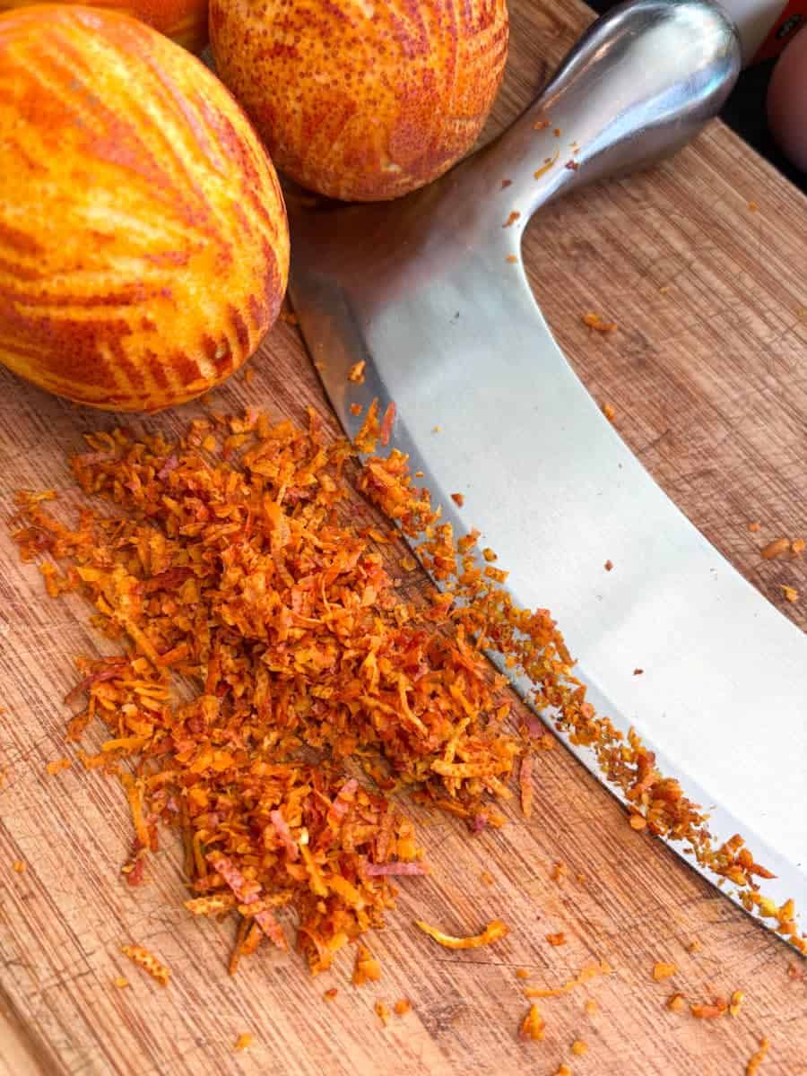 Grated orange zest and two oranges on a wooden chopping board with a silver mezzaluna.