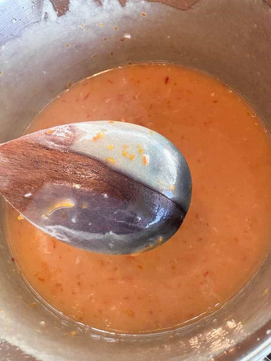 A silver pan of orange curd. A wooden spoon covered in curd is being lifted up to illustrate the curd has properly cooked.
