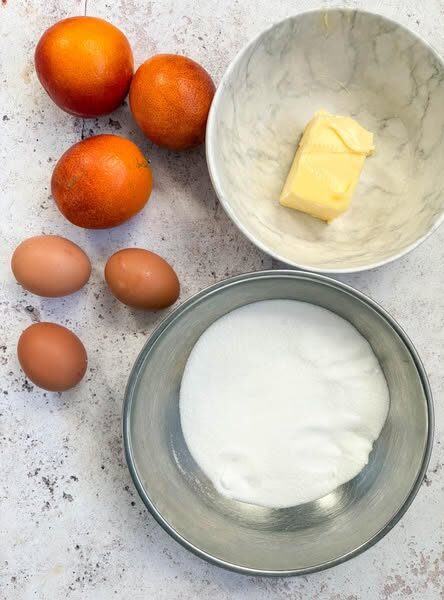 A silver bowl of white sugar, a white bowl of butter, three blood oranges and three eggs on a white and grey mottled surface.