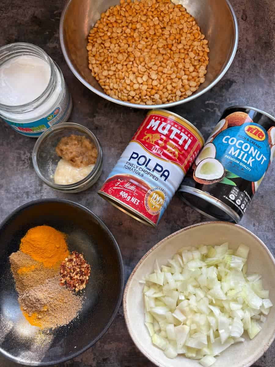 A silver bowl of yellow lentils, bowls of spices and chopped onion, a can of coconut milk, a can of chopped tomatoes, a glass dish of crushed garlic and ginger and a jar of coconut oil on a grey surface.