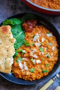 A black bowl of yellow lentil dahl sprinkled with fresh coriander and toasted coconut chips with a piece of Naan bread, fresh spinach leaves and a spoonful of mango chutney