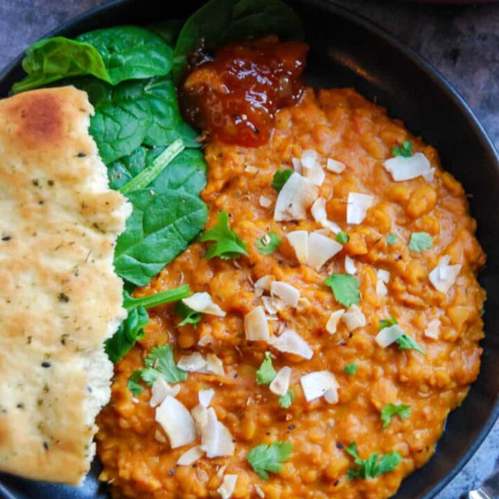 A black bowl of yellow lentil dahl sprinkled with fresh coriander and toasted coconut chips with a piece of Naan bread, fresh spinach leaves and a spoonful of mango chutney