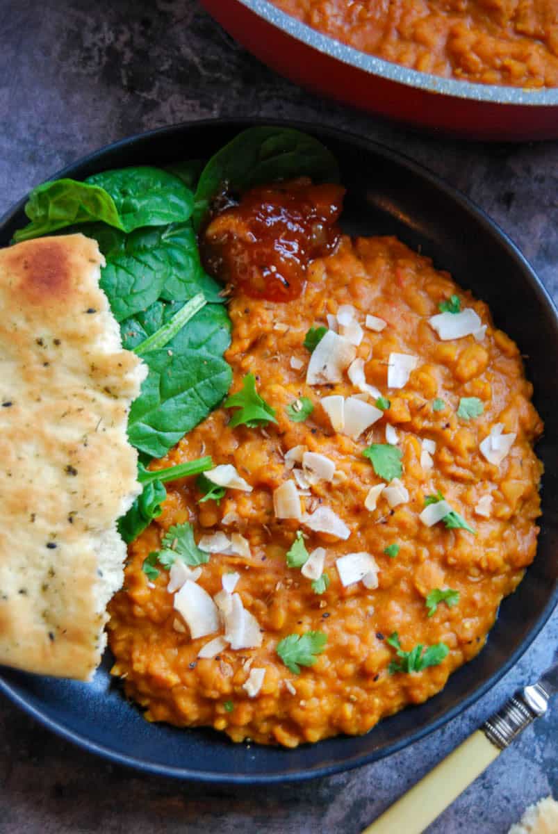 A black bowl of yellow lentil dahl sprinkled with fresh coriander and toasted coconut chips with a piece of Naan bread, fresh spinach leaves and a spoonful of mango chutney