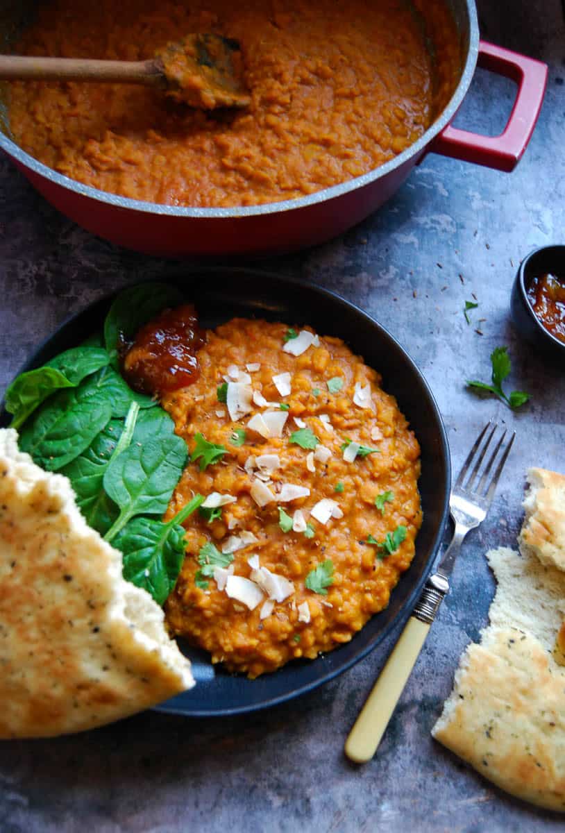 A black bowl of lentil dahl sprinkled with fresh coriander, toasted coconut chips, baby leaf spinach and a spoonful of mango chutney. A piece of Naan bread is propped against the bowl. A large red pan of dahl with a wooden spoon sits above the bowl.