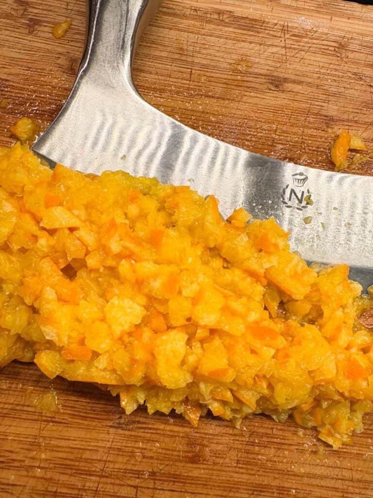 A cooked orange finely chopped into a rough puree on a wooden chopping board with a silver mezzaluna chopper.