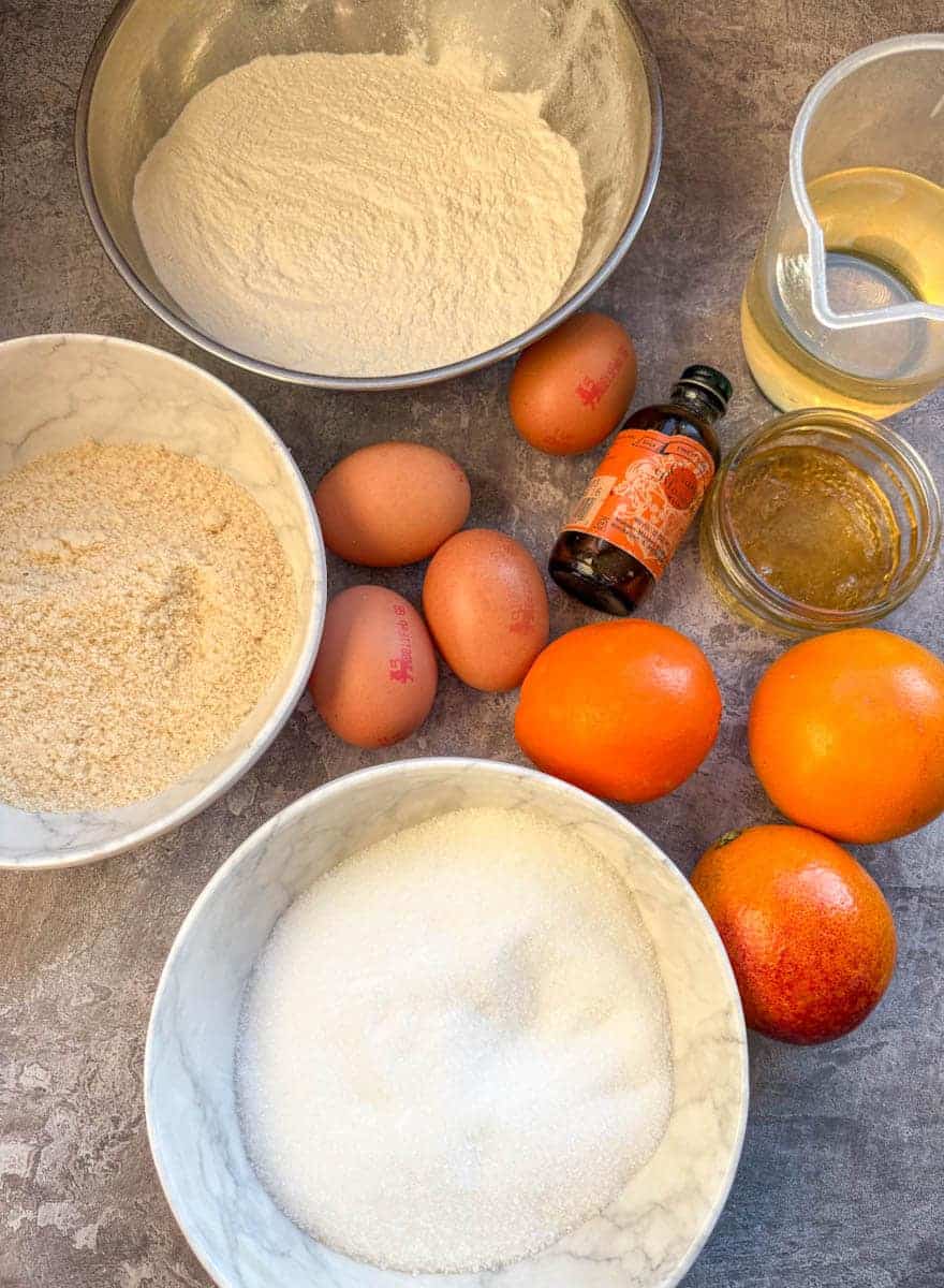 A silver bowl of flour, two white bowls of white sugar and ground almonds, a jug of oil, four eggs, three oranges, a glass dish of honey and a bottle of orange flower water.