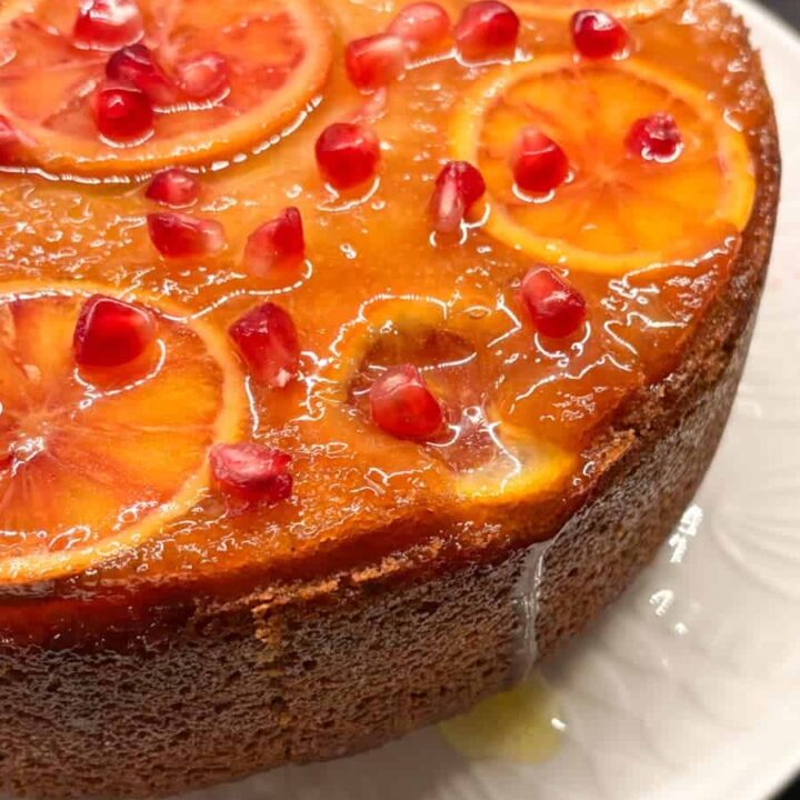 A upside down orange cake on a white plate. The top of the cake is adorned with sliced orange, pomegranate seeds and brushed with an orange syrup.