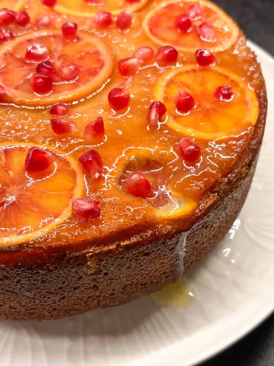 A upside down orange cake on a white plate. The top of the cake is adorned with sliced orange, pomegranate seeds and brushed with an orange syrup.