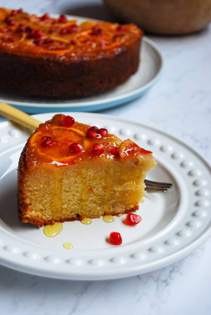 A slice of upside down orange cake on a white plate. The top of the cake is decorated with orange slices, pomegranate seeds and drizzled with an orange syrup that's running down the sides of the cake. A fork with a yellow handle is resting against the plate.