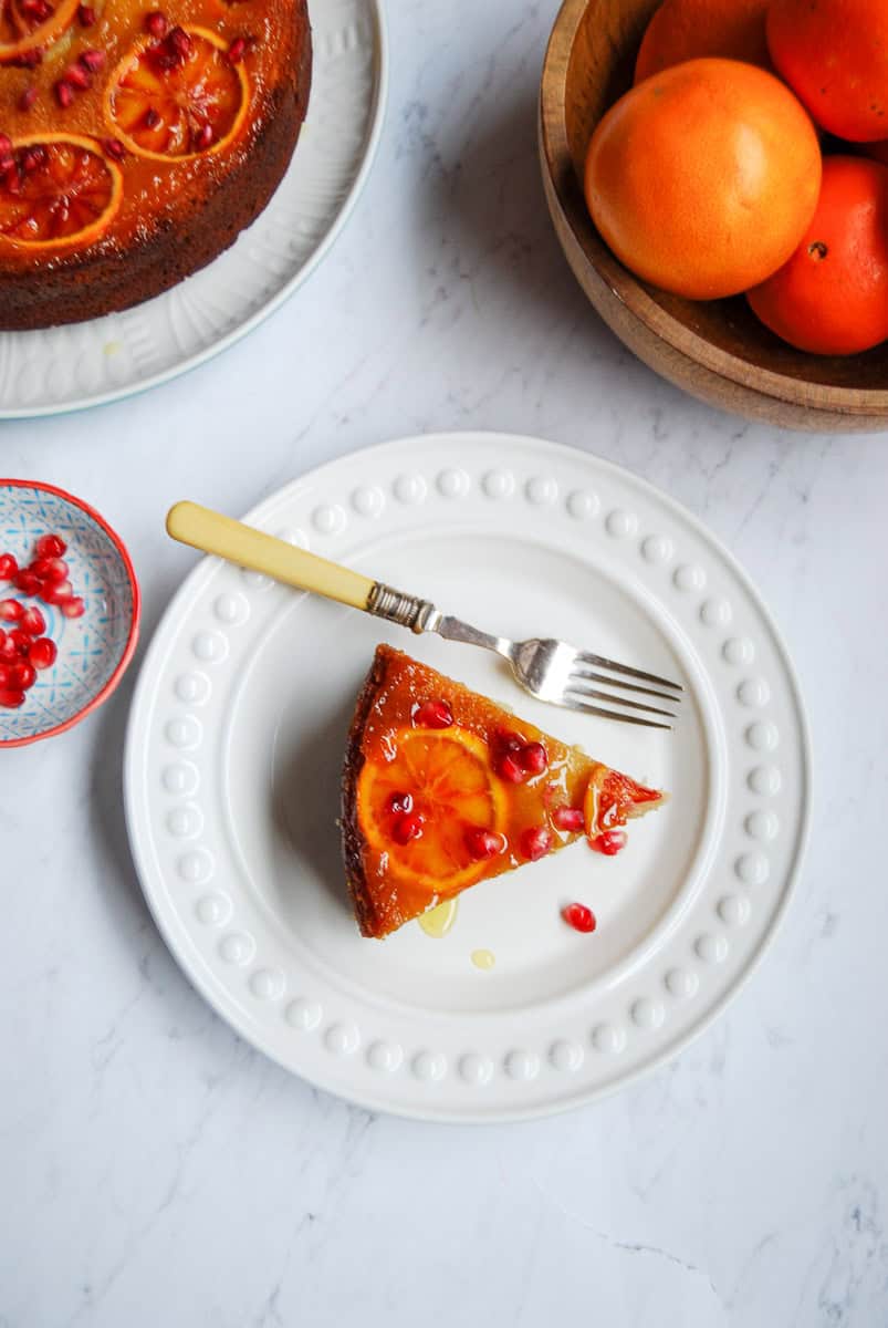 A slice of orange cake on a white plate. The cake is adorned with orange slices, pomegranate seeds and drizzled with orange syrup. A silver fork with a yellow handle is resting against the plate. A small dish of pomegranate seeds, a wooden bowl of oranges and an upside down orange cake sprinkled with pomegranate seeds.