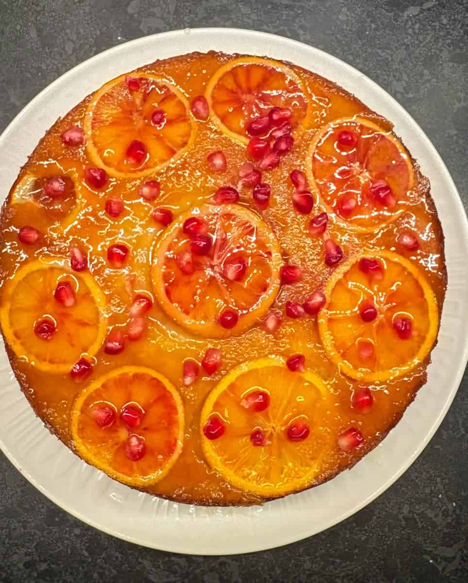 An upside down orange cake on a white plate. The top of the cake is decorated with sliced blood oranges, pomegranate seeds and brushed with orange syrup for a pretty shiny appearance.