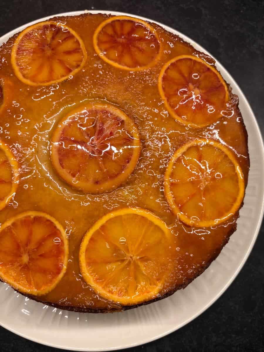 An upside down orange cake on a white plate. Sliced oranges adorn the top of the cake. Orange syrup has been brushed over the cake for an attractive glazed appearance.