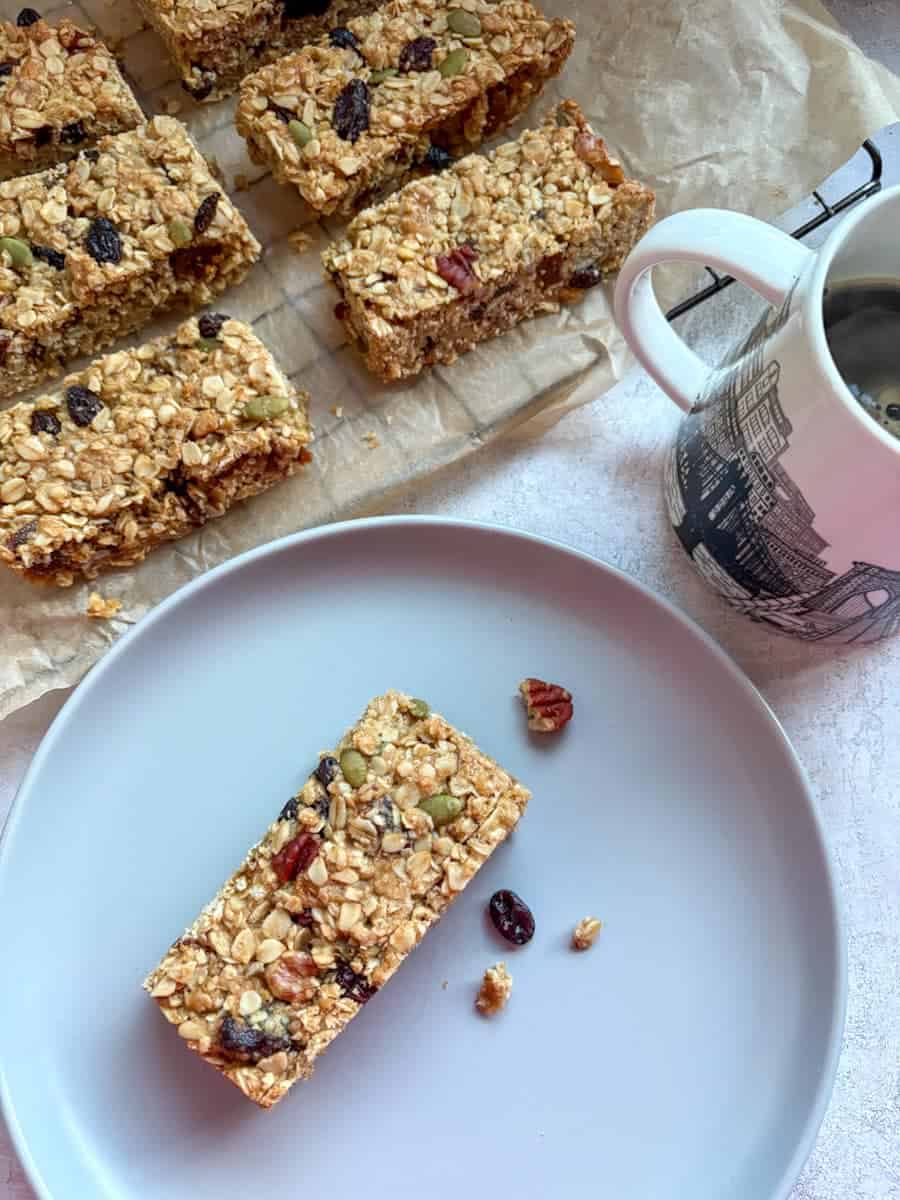 A granola bar on a blue plate, a white and black mug of coffee and a wire cooling rack lined with a piece of baking paper with more granola bars.