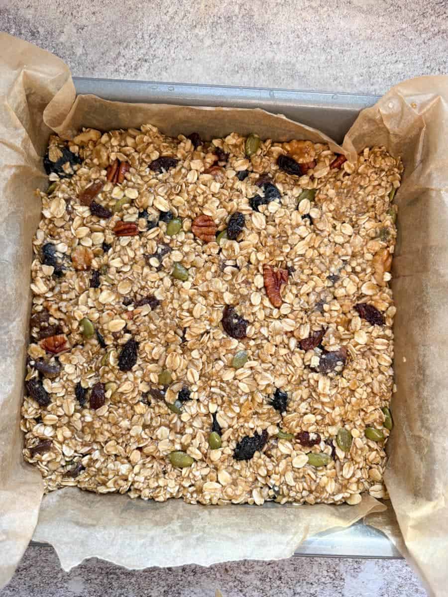 A silver pan lined with baking paper and granola bar mixture with chopped nuts, dried fruits and mixed seeds.