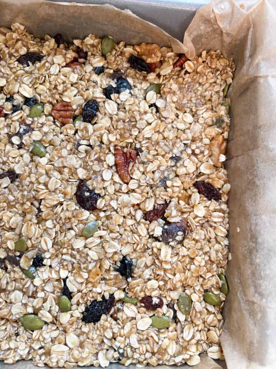 Unbaked granola bars with mixed seeds, chopped nuts and dried fruit inside a lined silver square baking tin.