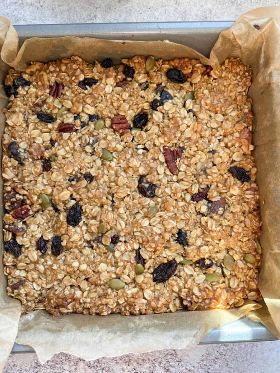 Freshly baked granola bars with mixed seeds, chopped nuts and dried fruit in a lined square baking tin.