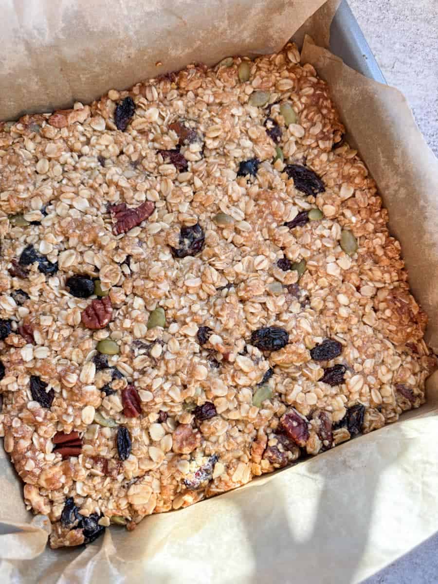 An unsliced granola bar with mixed nuts, dried fruit and seeds in a silver square baking tin lined with paper.