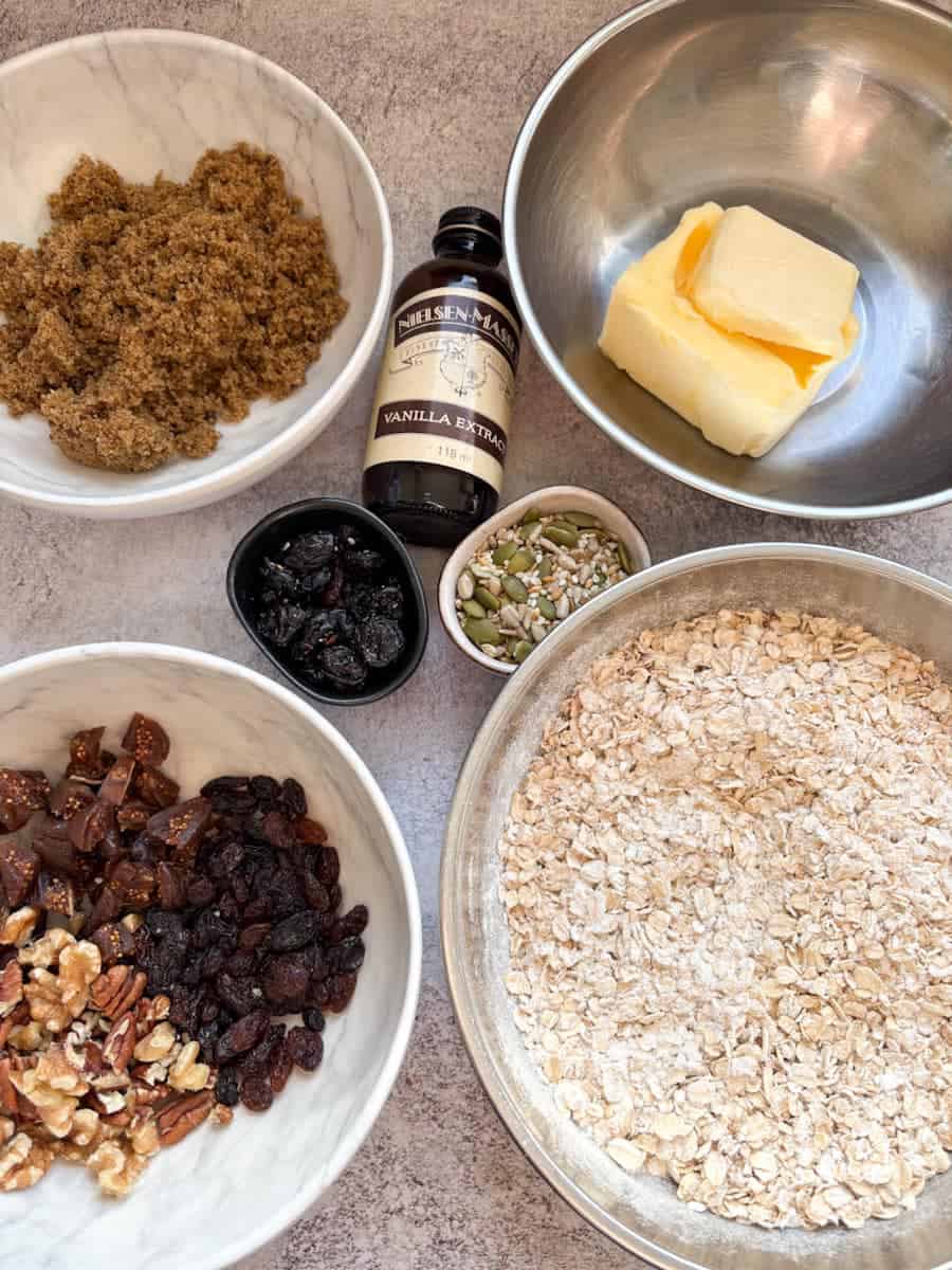 Bowls of various ingredients to make granola bars. Butter, brown sugar, oats, chopped nuts and dried fruits, a small pot of mixed seeds and a bottle of vanilla extract.